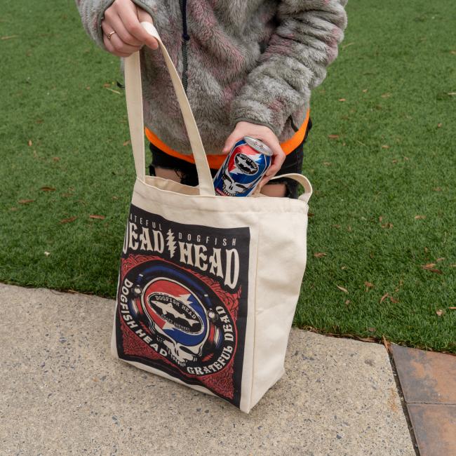 A person in a furry jacket stands on a sidewalk holding a can and the Dogfish Head x Grateful Dead Tote Bag by Red Promotions—unique craft beer merchandise displayed beside green grass.