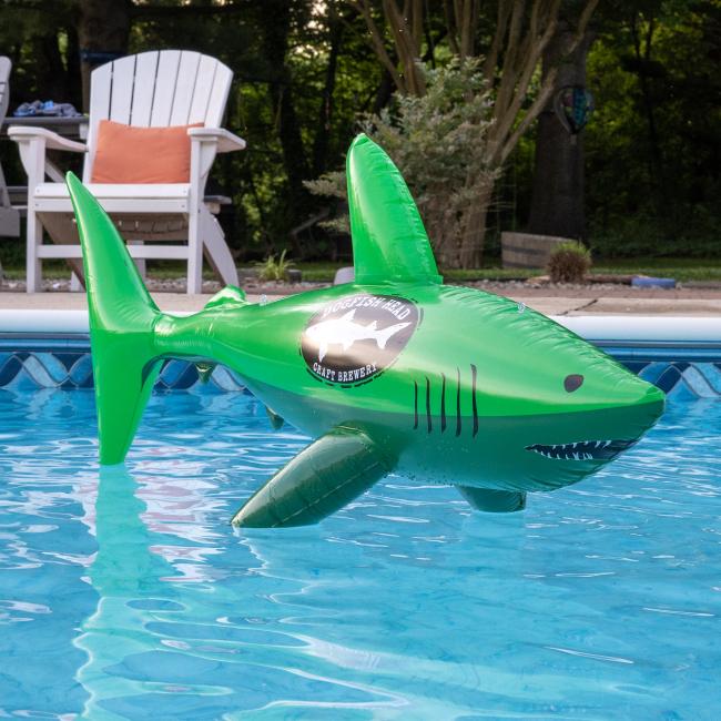 A Merchtank Dogfish Pool Float with a shiny logo floats in a pool, surrounded by white chairs and trees.