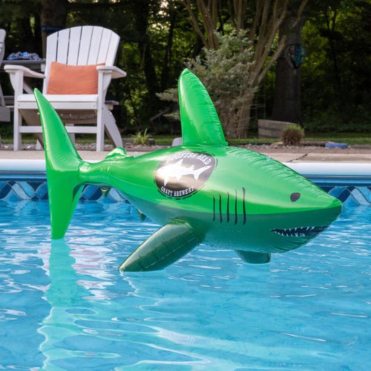 A Merchtank Dogfish Pool Float with a shiny logo floats in a pool, surrounded by white chairs and trees.
