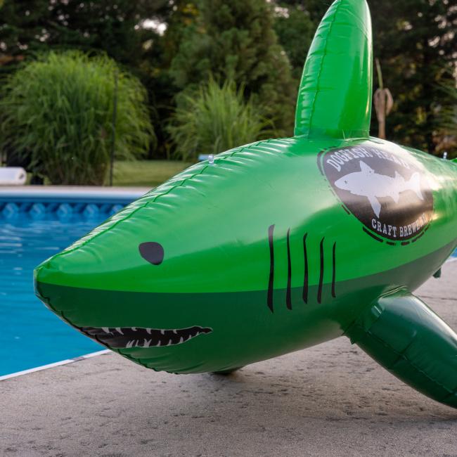 The Merchtank Dogfish Pool Float, featuring a green vinyl design with a brewery logo, sits on the poolside amid lush greenery and trees.