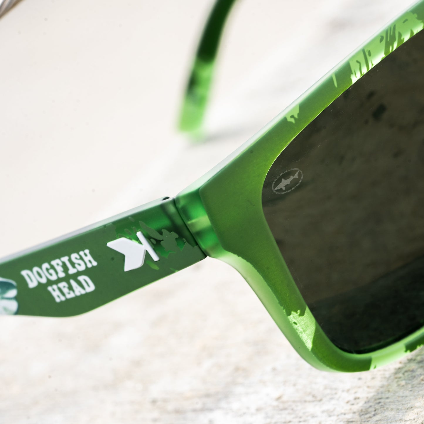 Close-up of green sunglasses with dark lenses and a green temple arm printed with the text 'DOGFISH HEAD' and a white fish logo, resting on a light textured surface.