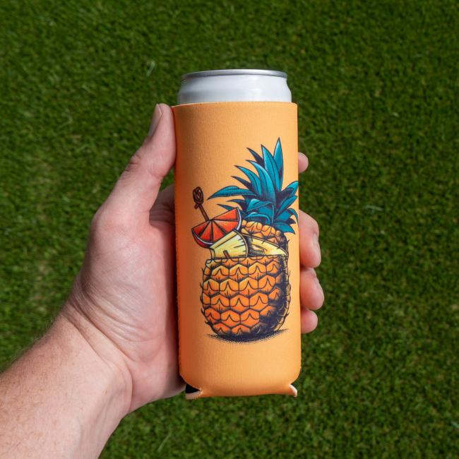 A hand holds a Merchtank Four Slim Can Cooler Bundle sleeve, orange with a vibrant pineapple, drink umbrella, and citrus slice illustration. The 12oz can is shown over green grass.