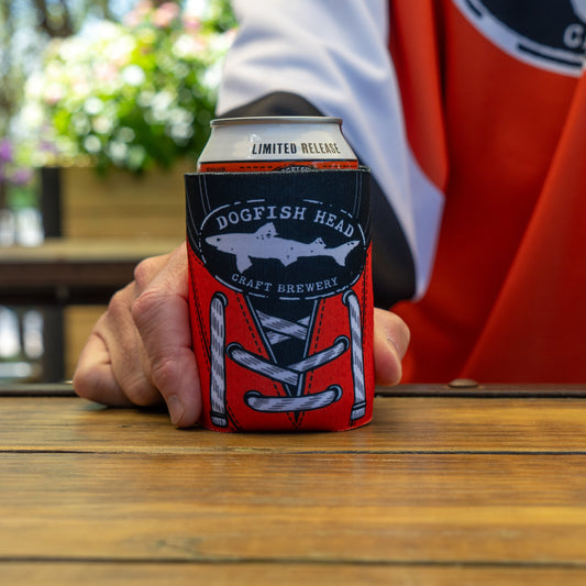 A person in an orange jersey holds a Dogfish Head x Garnet Hathaway Engine 19 12oz Can Cooler by Grandstand Glassware & Apparel, resting on a wooden table with blurred outdoor greenery in the background.