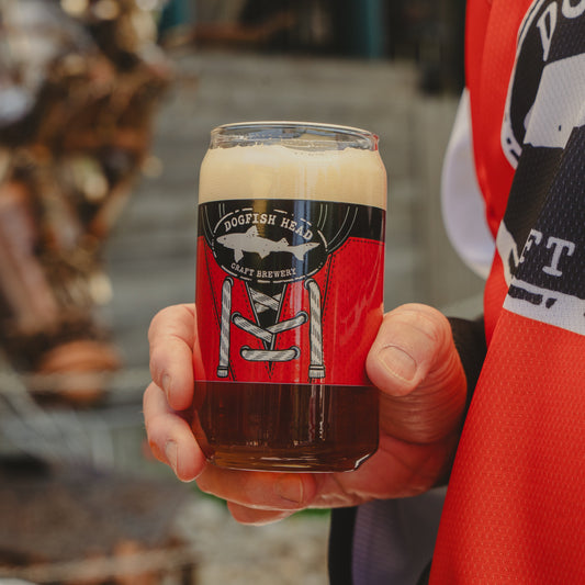 A hand holds the Dogfish Head x Garnet Hathaway Engine 19 Can Glass by Grandstand Glassware & Apparel, styled like a red hockey jersey with laces and engine branding, while the person wears a matching Hath's Heroes jersey.