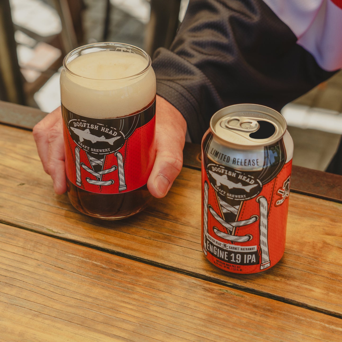 A person holds a glass of beer next to a Dogfish Head x Garnet Hathaway Engine 19 Can Glass by Grandstand Glassware & Apparel on a wooden table. Both feature red and black football jersey-inspired designs celebrating their collaboration.
