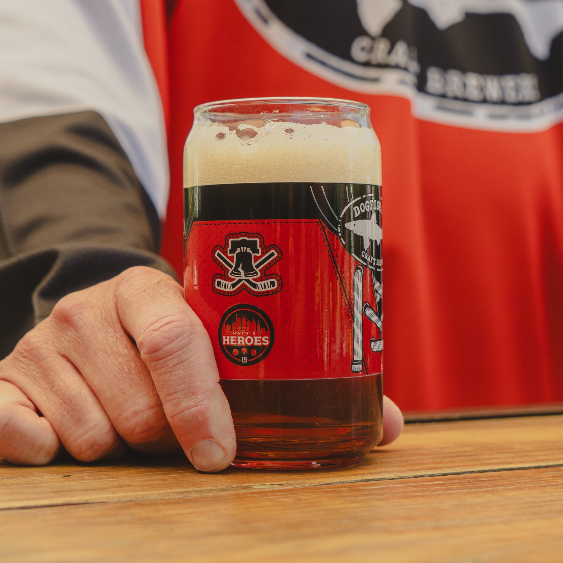 A person in a red and black shirt holds a Dogfish Head x Garnet Hathaway Engine 19 Can Glass by Grandstand Glassware & Apparel, filled with dark beer and foam, on a wooden table.