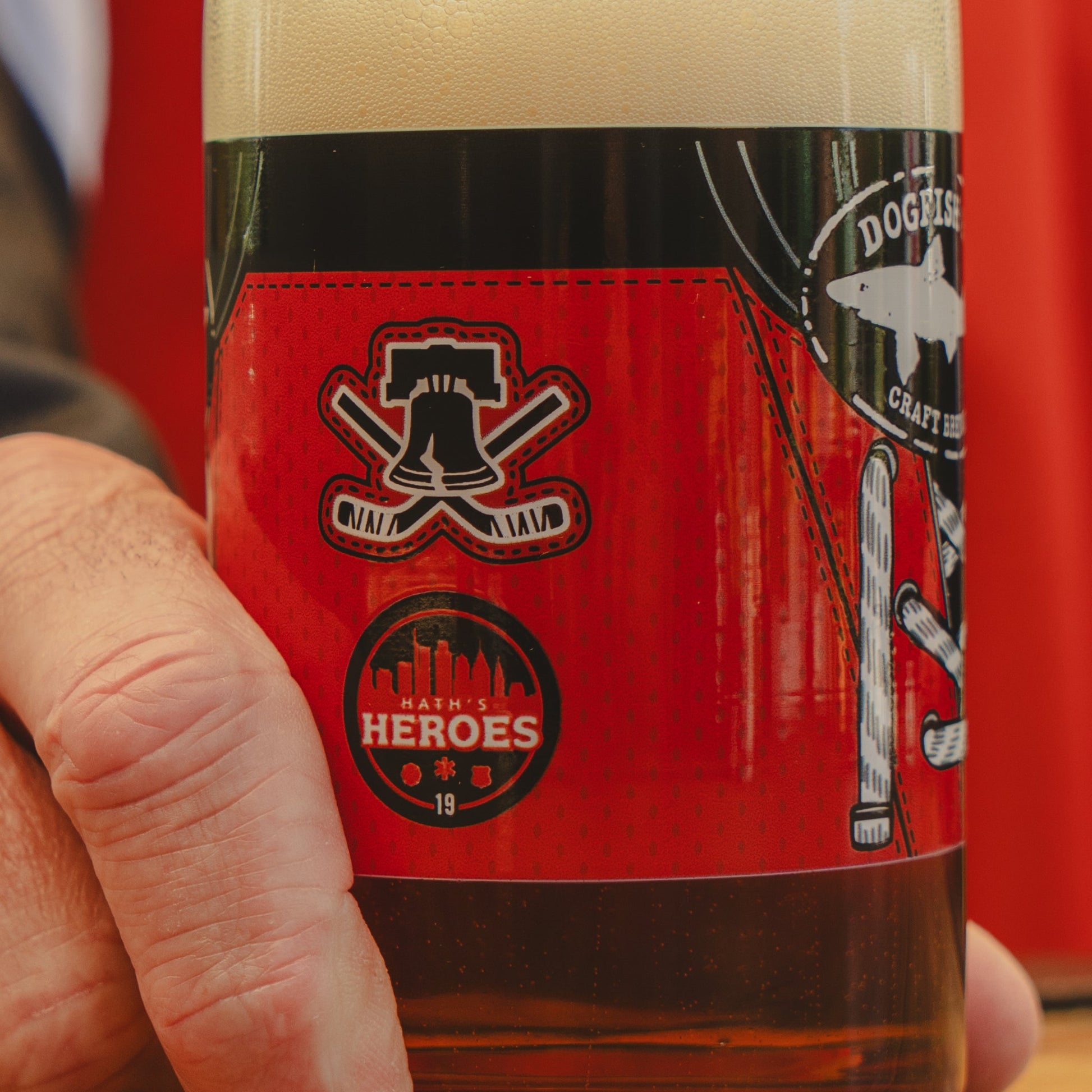 A close-up of a hand holding the Dogfish Head x Garnet Hathaway Engine 19 Can Glass by Grandstand Glassware & Apparel, featuring a city skyline, bell, crossed bats, and the words "Hath's Heroes" and "Engine 19 IPA.