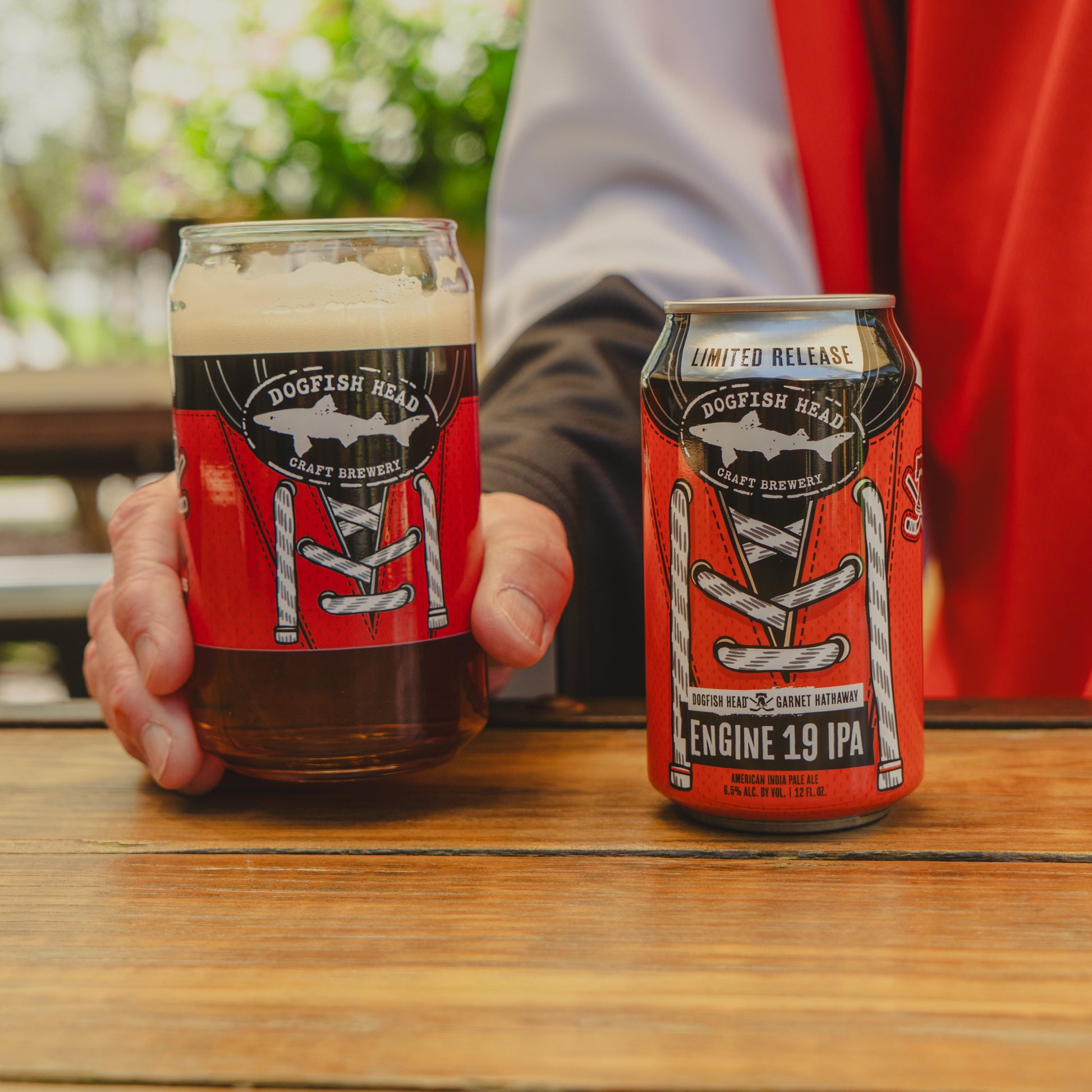 A person holds a Dogfish Head x Garnet Hathaway Engine 19 Can Glass and a matching can with a fireman’s jacket red label, both from Grandstand Glassware & Apparel, resting on a wooden outdoor table.