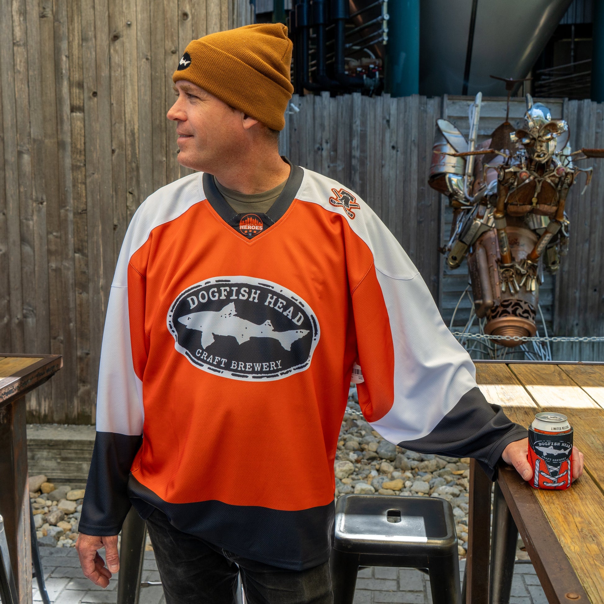 A man in a Dogfish Head x Garnet Hathaway Engine 19 Hockey Jersey by In Your Face Apparel and a brown beanie stands outdoors by a table, holding a can, with a wooden fence and metal humanoid sculpture behind him.