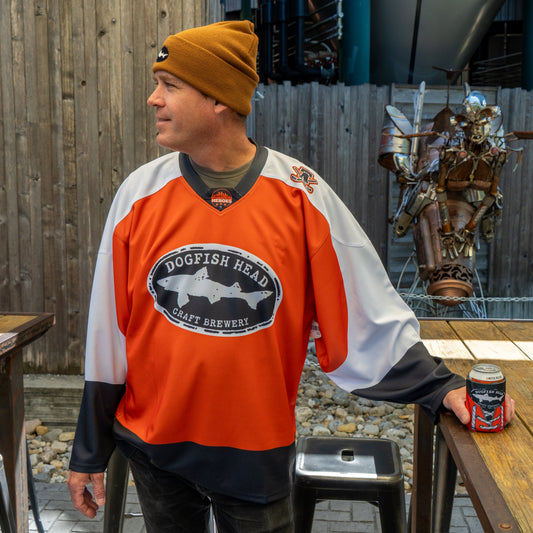A man in a Dogfish Head x Garnet Hathaway Engine 19 Hockey Jersey by In Your Face Apparel and a brown beanie stands outdoors by a table, holding a can, with a wooden fence and metal humanoid sculpture behind him.