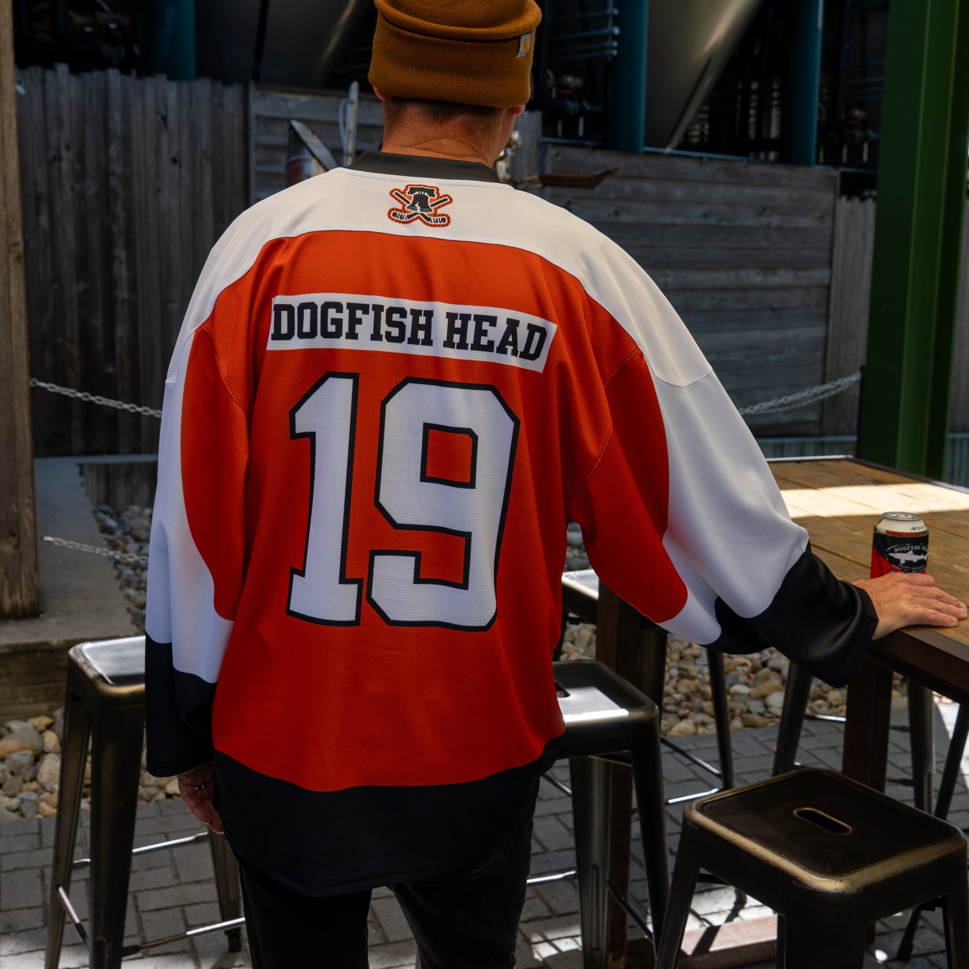 Wearing an In Your Face Apparel Dogfish Head x Garnet Hathaway Engine 19 Hockey Jersey, a person in a brown beanie stands by a metal bar table outdoors, holding a can, celebrating first responders.