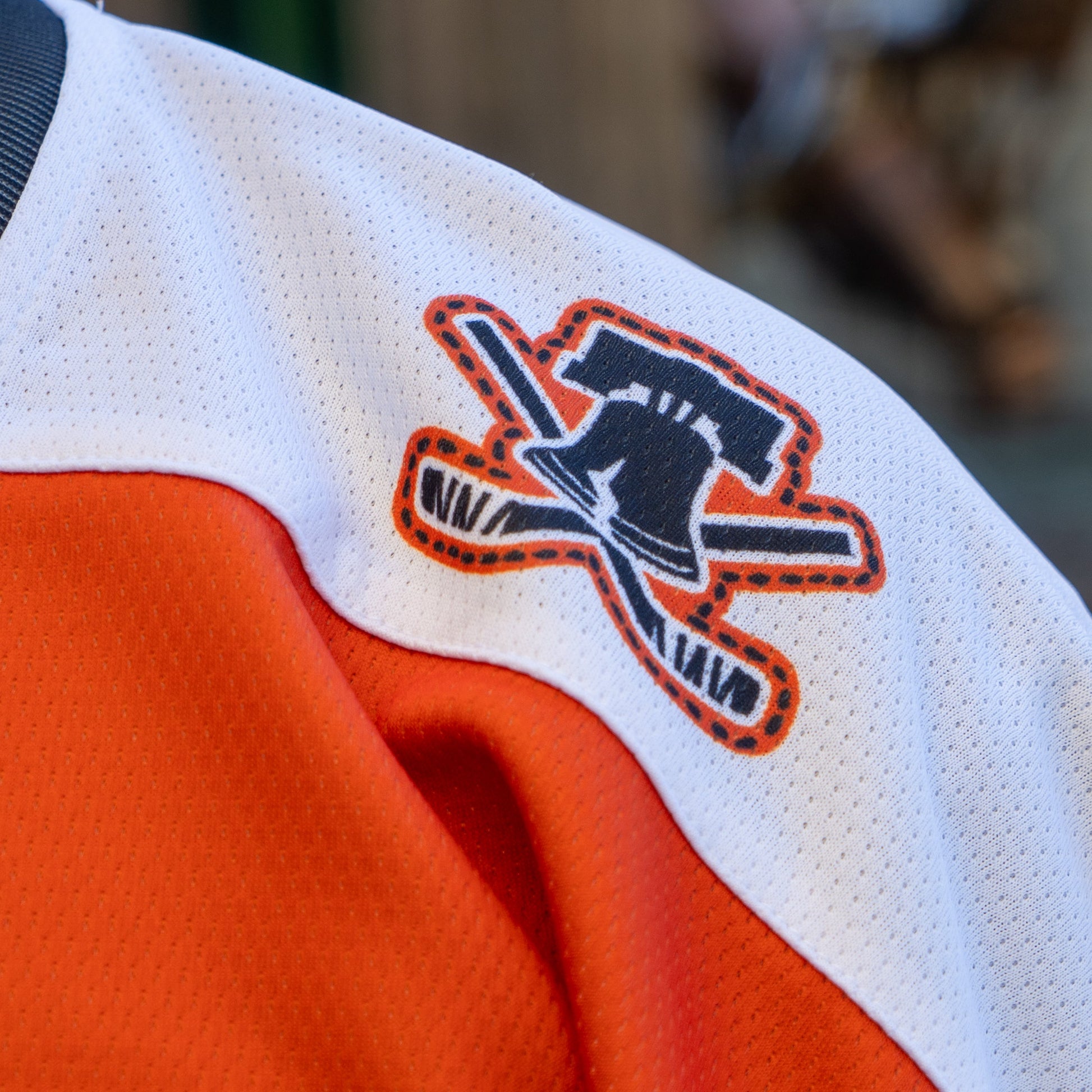 Close-up of the Dogfish Head x Garnet Hathaway Engine 19 Hockey Jersey by In Your Face Apparel, showing a shoulder patch with a black Liberty Bell and orange hockey sticks on white—boldly honoring first responders in orange and white.