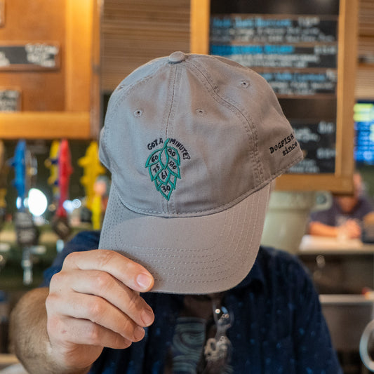 A person holds the Got A Minute? Dad Hat by L2 Brands in front of their face, showing off the Minute Series hop logo and "Got a minute? Dogfish Head since 1995" text against a bar backdrop with a chalkboard menu.