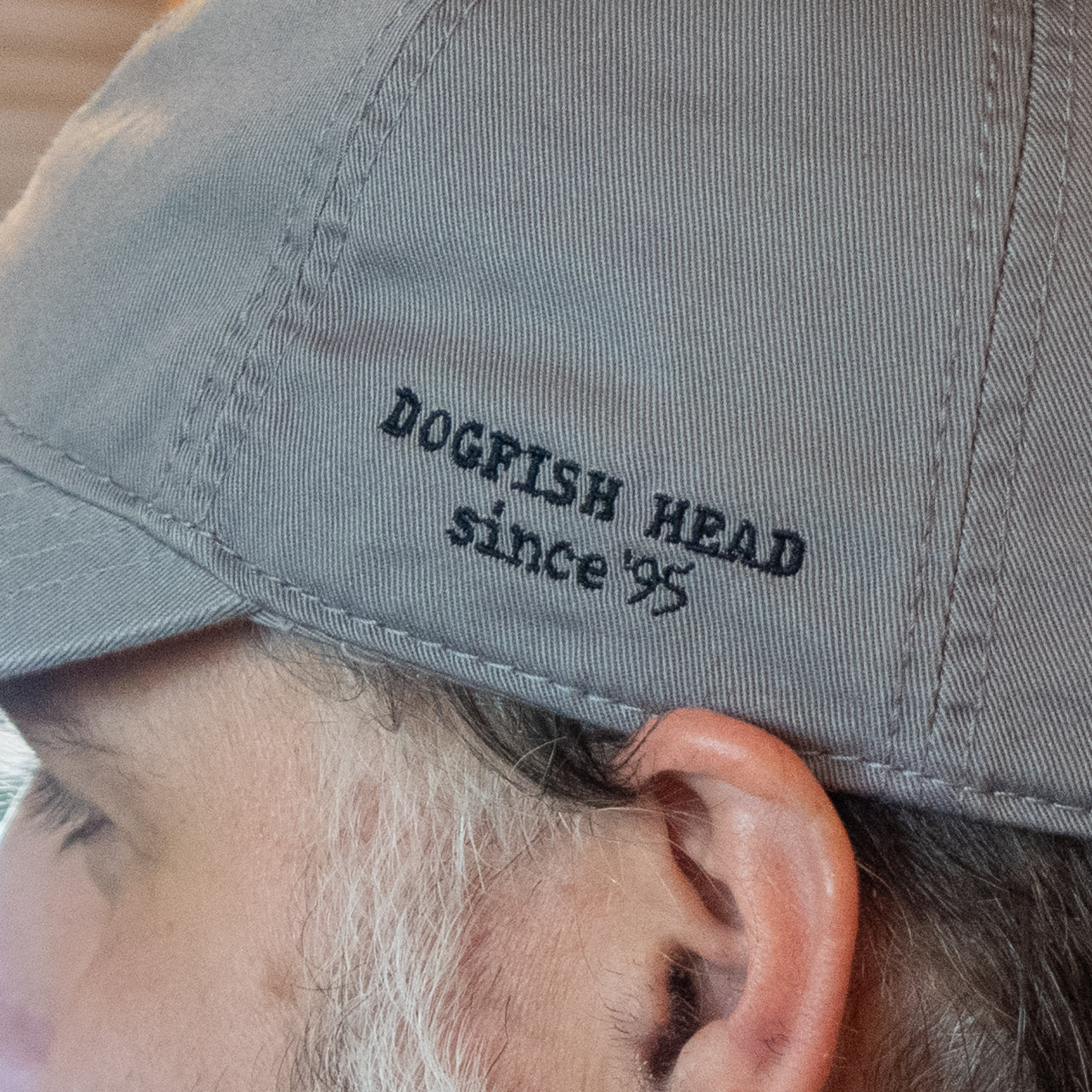 Close-up of a person wearing the Got A Minute? Dad Hat by L2 Brands, a gray cap with black embroidered text "DOGFISH HEAD since 95" and a subtle hop logo that hints at their signature hop-forward style.