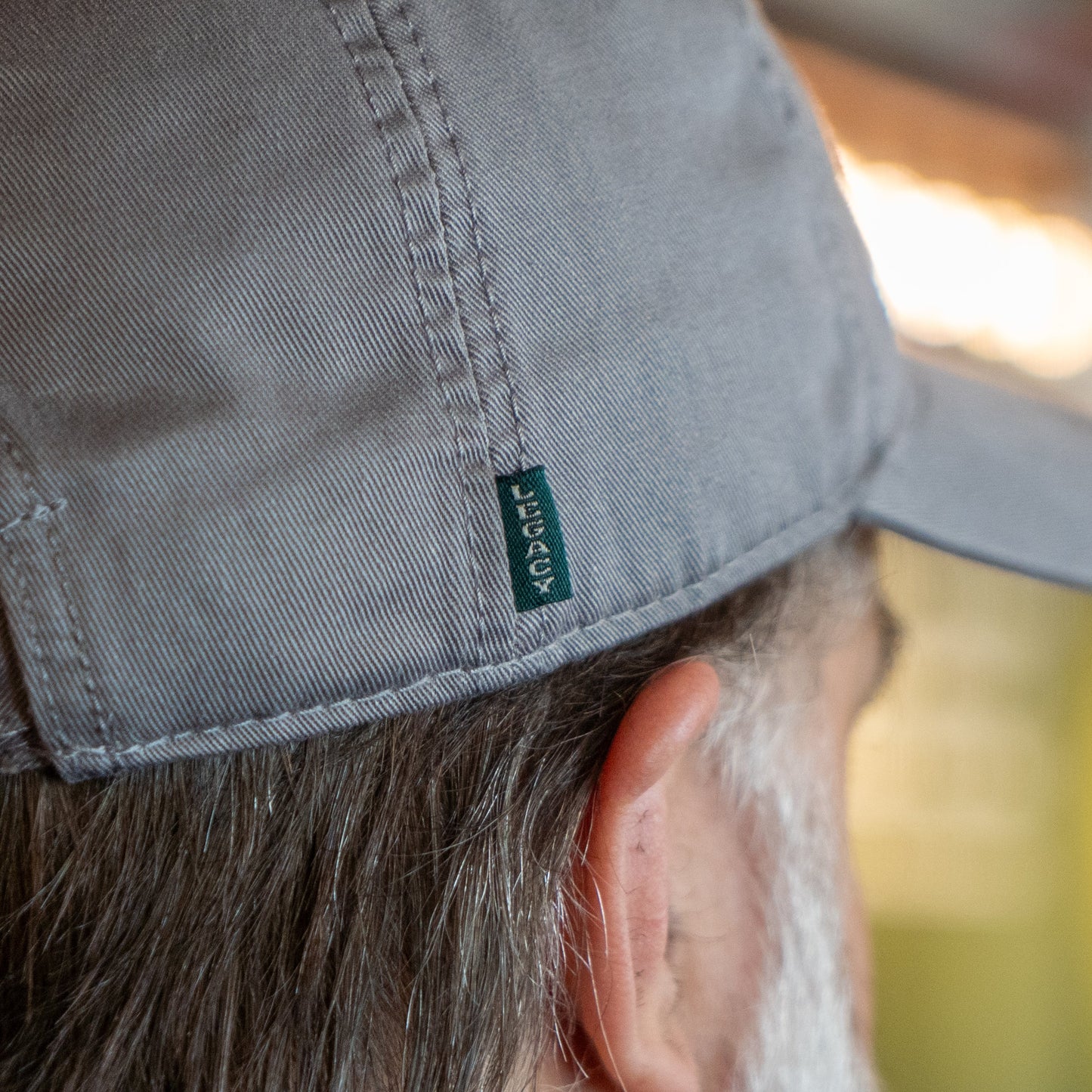 Close-up of the side and back of a person's head wearing the Got A Minute? Dad Hat by L2 Brands in light gray, featuring a small green LEGACY tag and the hop logo from the Minute Series. The person's ear and gray hair are partially visible; background is blurred.