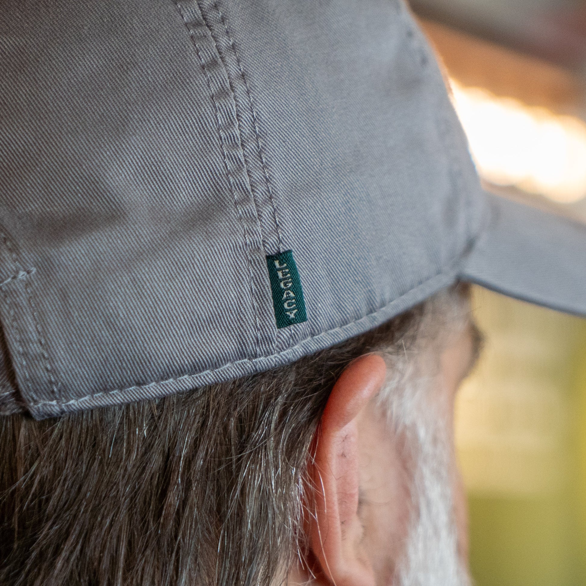 Close-up of the side and back of a person's head wearing the Got A Minute? Dad Hat by L2 Brands in light gray, featuring a small green LEGACY tag and the hop logo from the Minute Series. The person's ear and gray hair are partially visible; background is blurred.
