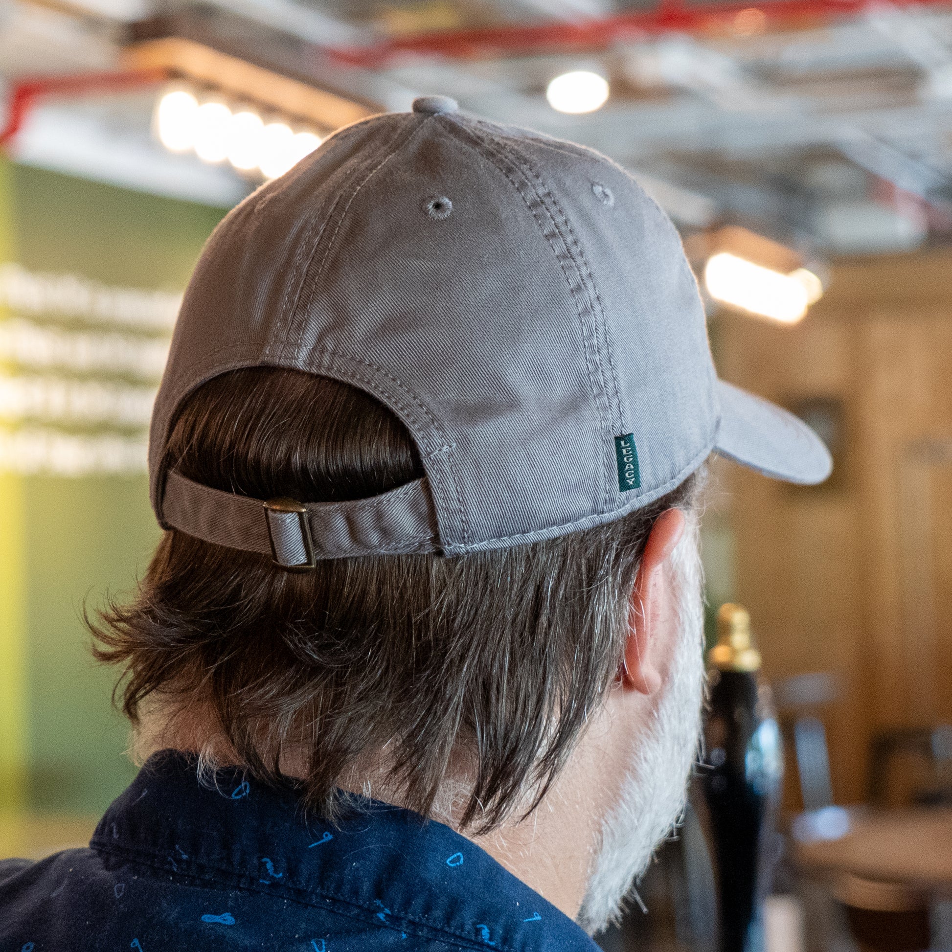 A man is seen from behind indoors, wearing the L2 Brands "Got A Minute? Dad Hat" in gray, featuring a subtle hop logo, along with a navy blue shirt. Blurred lights and a green wall are visible in the background.