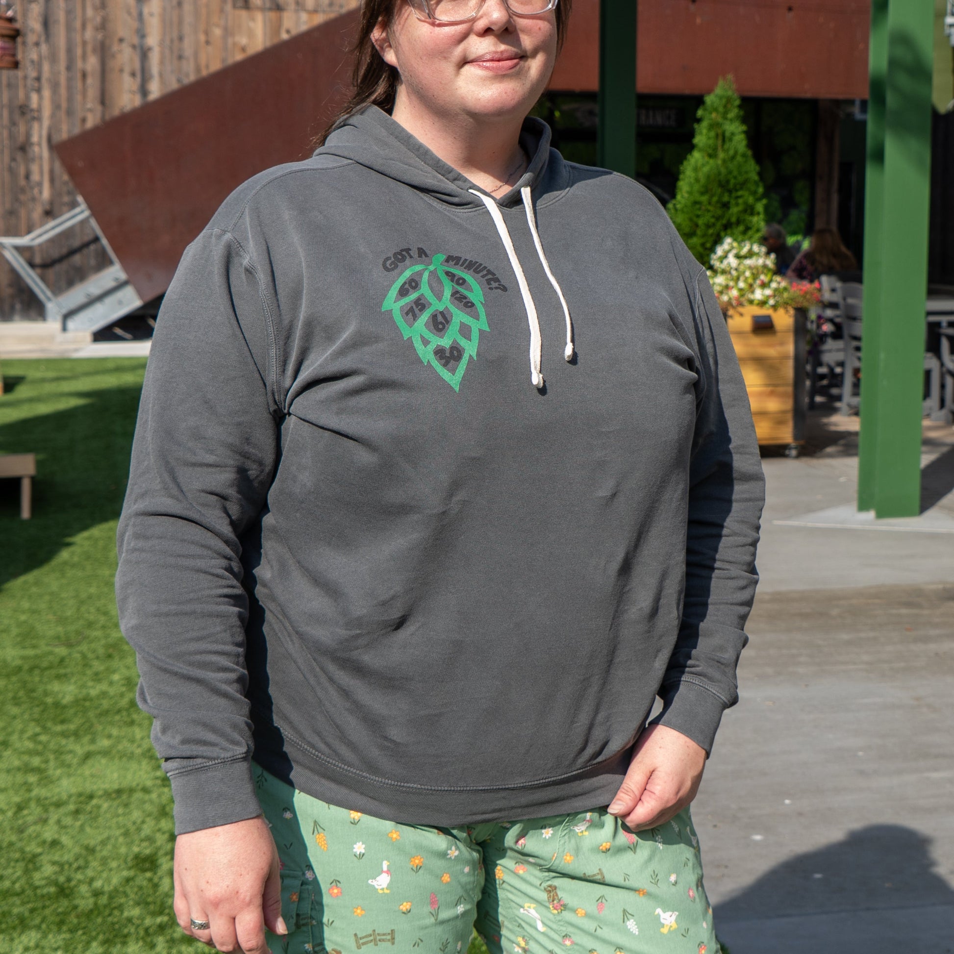 A person wearing a gray GO WEST T-SHIRT COMPANY Got a Minute? Hoodie with a bold hop logo and light green pajama pants with small animal patterns stands outside on a sunny day, with buildings and greenery behind them.
