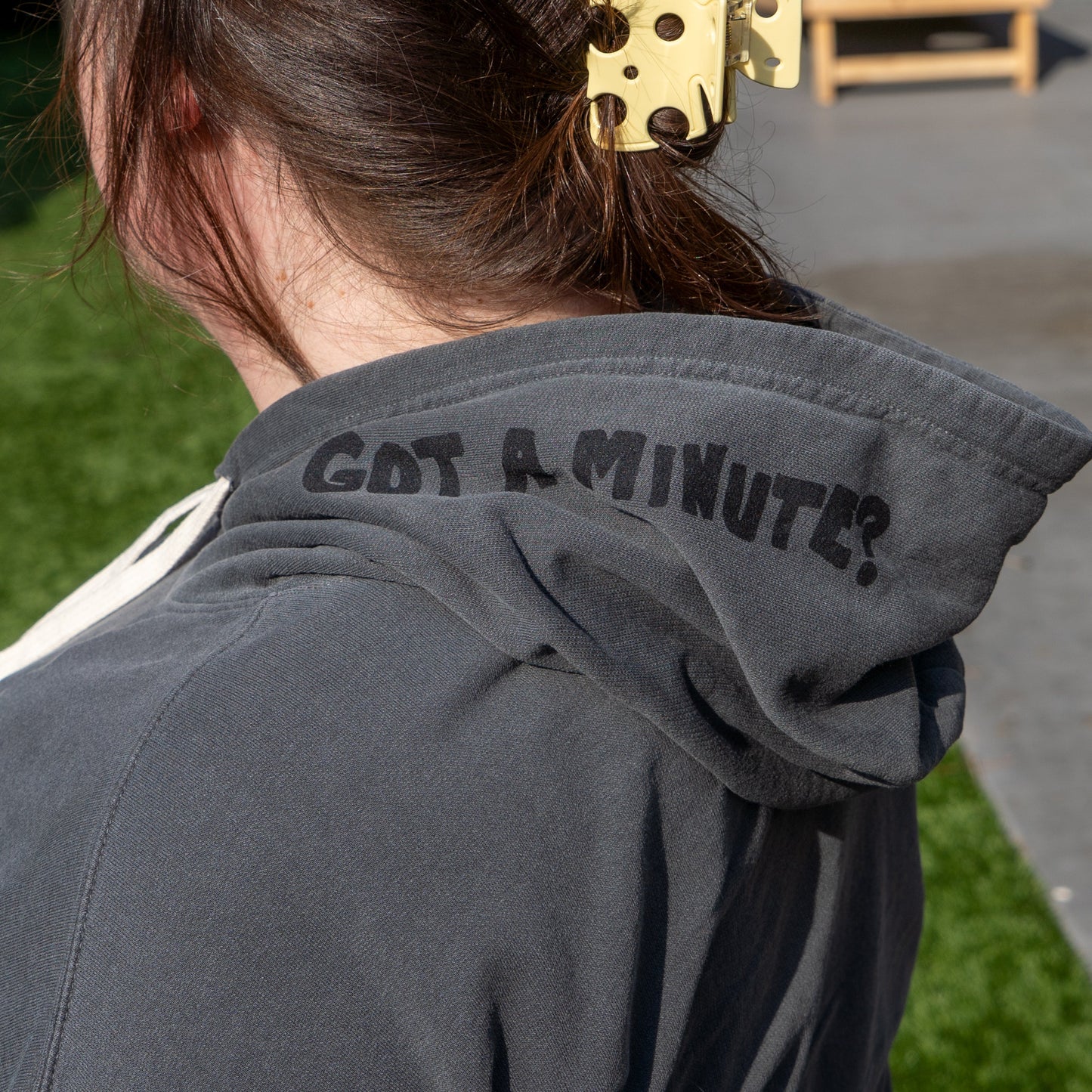 A person with brown hair in a yellow clip sports the gray Got a Minute? Hoodie from GO WEST T-SHIRT COMPANY, featuring “GOT A MINUTE?” on the hood, outside on a sunny day—ideal for showcasing this bold style.