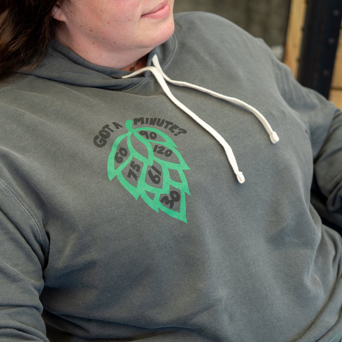 A person wears the Got a Minute? Hoodie by GO WEST T-SHIRT COMPANY—a gray hoodie with white drawstrings and a bold hop logo featuring clock numbers, topped by the text GOT A MINUTE?.