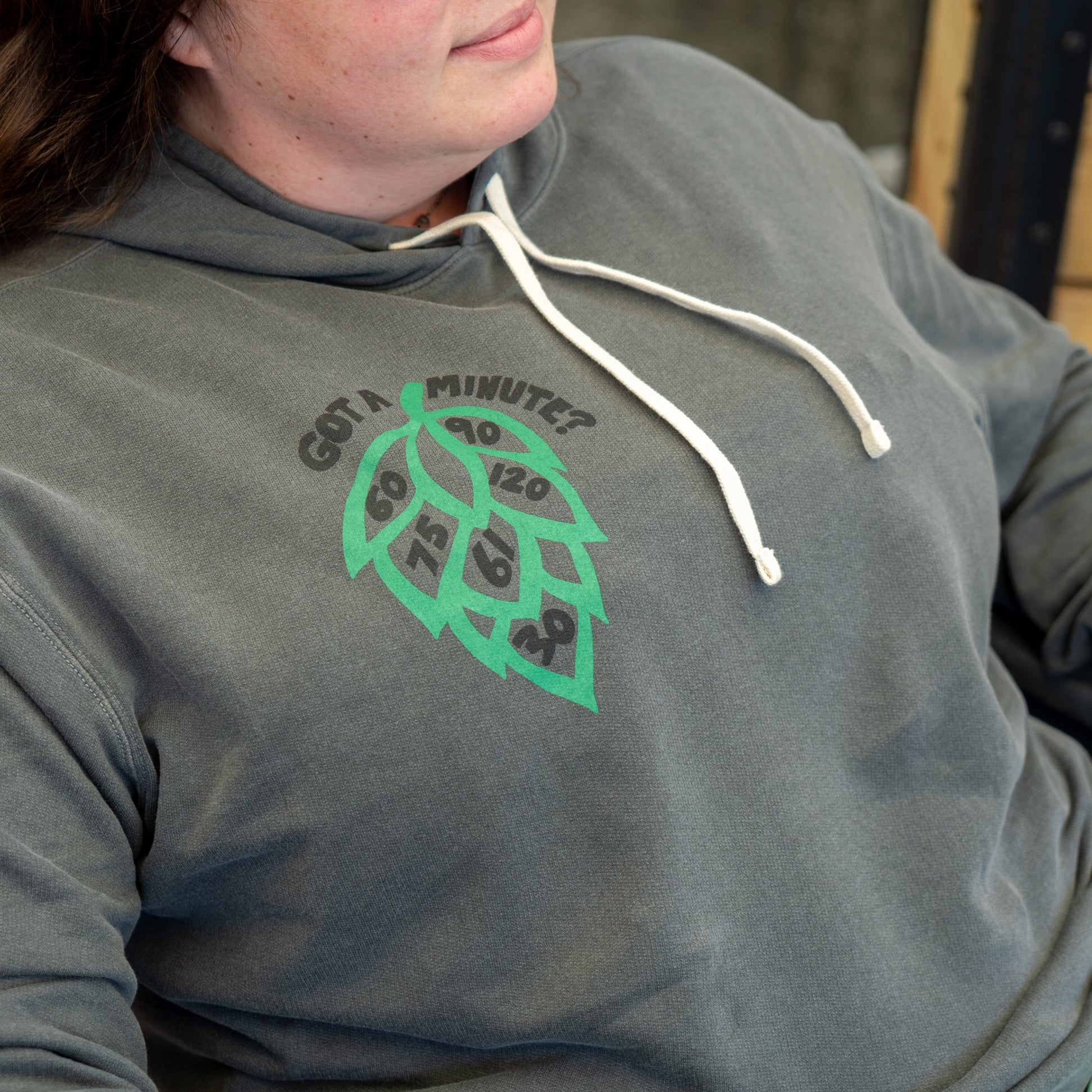 A person wears the Got a Minute? Hoodie by GO WEST T-SHIRT COMPANY—a gray hoodie with white drawstrings and a bold hop logo featuring clock numbers, topped by the text GOT A MINUTE?.