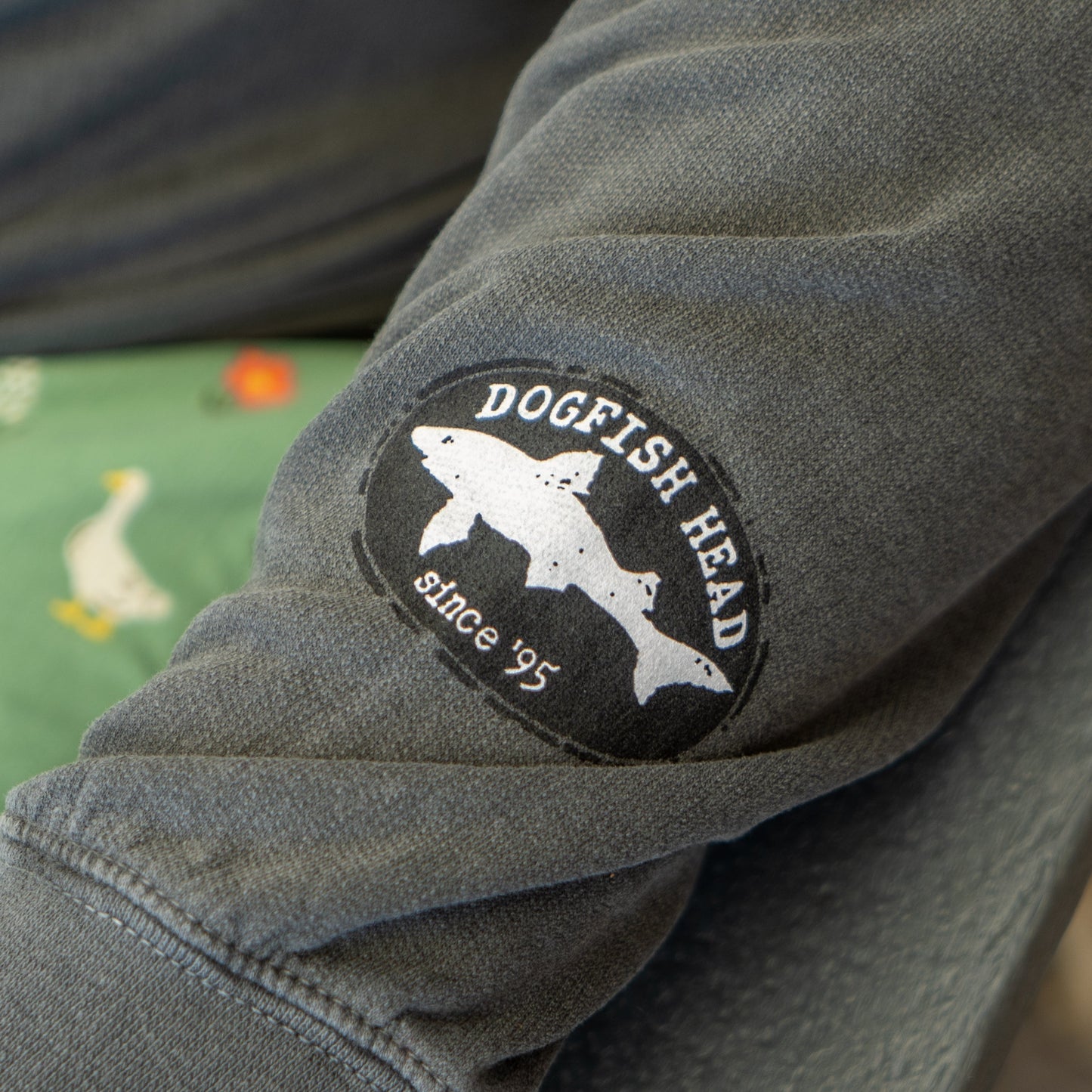 A close-up of a gray Got a Minute? Hoodie sleeve by GO WEST T-SHIRT COMPANY, featuring a circular patch with three overlapping fish and "Dogfish Head since 95." In the background, green fabric with white ducks is partially visible.