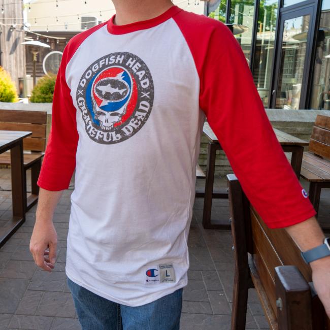 White three quarter sleeve tee with red sleeves. Tee has a black emblem on the center with "Dogfish Head" "Grateful Dead" written around the circle. White skull is featured in the center wearing blue and red headphones. In the head of the skull is a black Dogfish Head shark and shield logo with the background of a circle which is half red and half blue with a white lightning bolt going through it. Champion logo featured on the bottom of the left sleeve and on the bottom left of the body of the tee.
