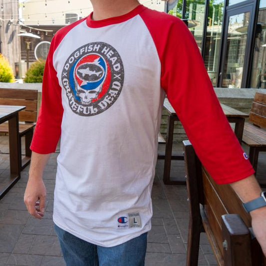 White three quarter sleeve tee with red sleeves. Tee has a black emblem on the center with "Dogfish Head" "Grateful Dead" written around the circle. White skull is featured in the center wearing blue and red headphones. In the head of the skull is a black Dogfish Head shark and shield logo with the background of a circle which is half red and half blue with a white lightning bolt going through it. Champion logo featured on the bottom of the left sleeve and on the bottom left of the body of the tee.