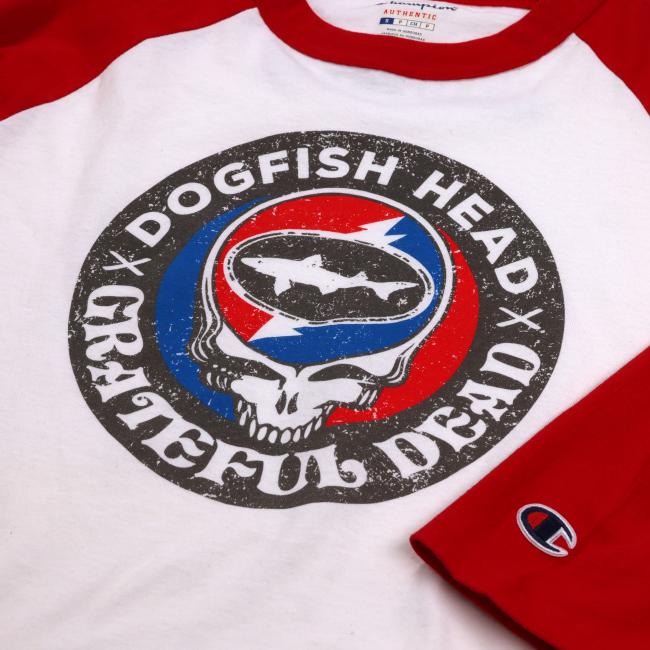 White three quarter sleeve tee with red sleeves. Tee has a black emblem on the center with "Dogfish Head" "Grateful Dead" written around the circle. White skull is featured in the center wearing blue and red headphones. In the head of the skull is a black Dogfish Head shark and shield logo with the background of a circle which is half red and half blue with a white lightning bolt going through it. Champion logo featured on the bottom of the left sleeve and on the bottom left of the body of the tee.