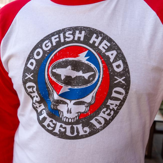 White three quarter sleeve tee with red sleeves. Tee has a black emblem on the center with "Dogfish Head" "Grateful Dead" written around the circle. White skull is featured in the center wearing blue and red headphones. In the head of the skull is a black Dogfish Head shark and shield logo with the background of a circle which is half red and half blue with a white lightning bolt going through it. Champion logo featured on the bottom of the left sleeve and on the bottom left of the body of the tee.