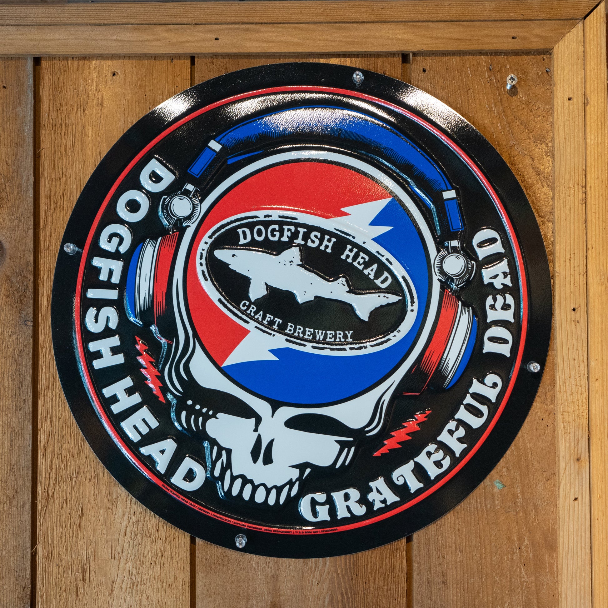 Black round tin tacker with "Dogfish Head" "Grateful Dead" written around the circle. White skull is featured in the center wearing blue and red headphones. In the head of the skull is a black Dogfish Head shark and shield logo with the background of a circle which is half red and half blue with a white lightning bolt going through it.