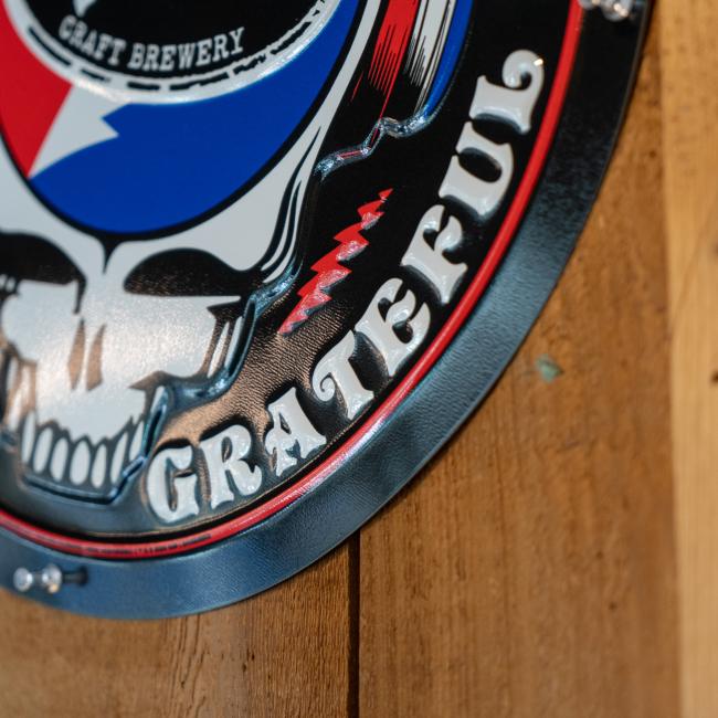 Black round tin tacker with "Dogfish Head" "Grateful Dead" written around the circle. White skull is featured in the center wearing blue and red headphones. In the head of the skull is a black Dogfish Head shark and shield logo with the background of a circle which is half red and half blue with a white lightning bolt going through it.