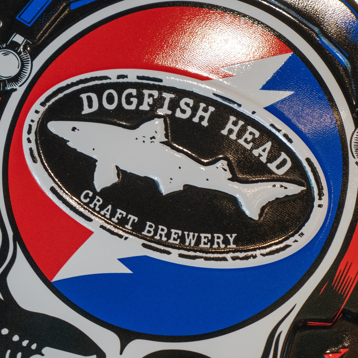 Black round tin tacker with "Dogfish Head" "Grateful Dead" written around the circle. White skull is featured in the center wearing blue and red headphones. In the head of the skull is a black Dogfish Head shark and shield logo with the background of a circle which is half red and half blue with a white lightning bolt going through it.