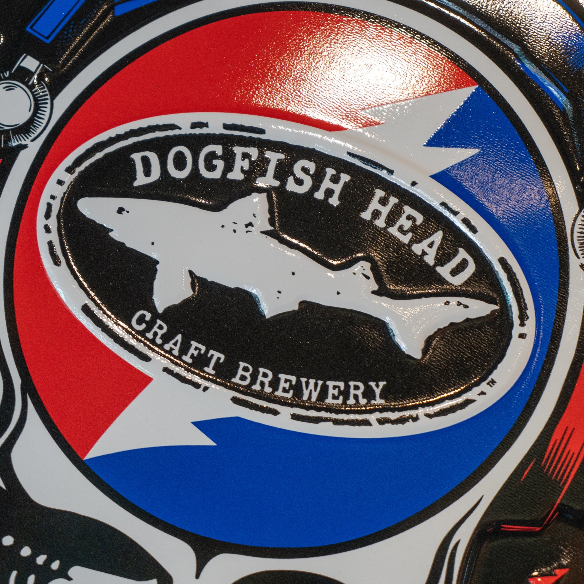 Black round tin tacker with "Dogfish Head" "Grateful Dead" written around the circle. White skull is featured in the center wearing blue and red headphones. In the head of the skull is a black Dogfish Head shark and shield logo with the background of a circle which is half red and half blue with a white lightning bolt going through it.
