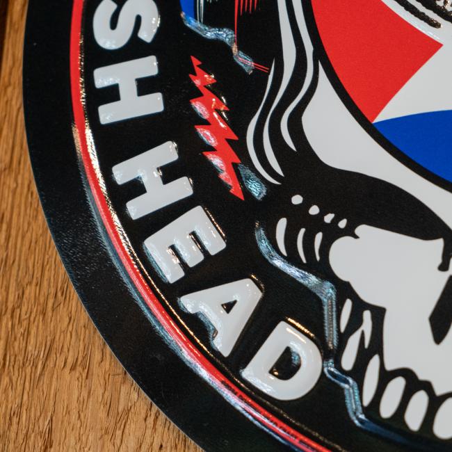 Black round tin tacker with "Dogfish Head" "Grateful Dead" written around the circle. White skull is featured in the center wearing blue and red headphones. In the head of the skull is a black Dogfish Head shark and shield logo with the background of a circle which is half red and half blue with a white lightning bolt going through it.