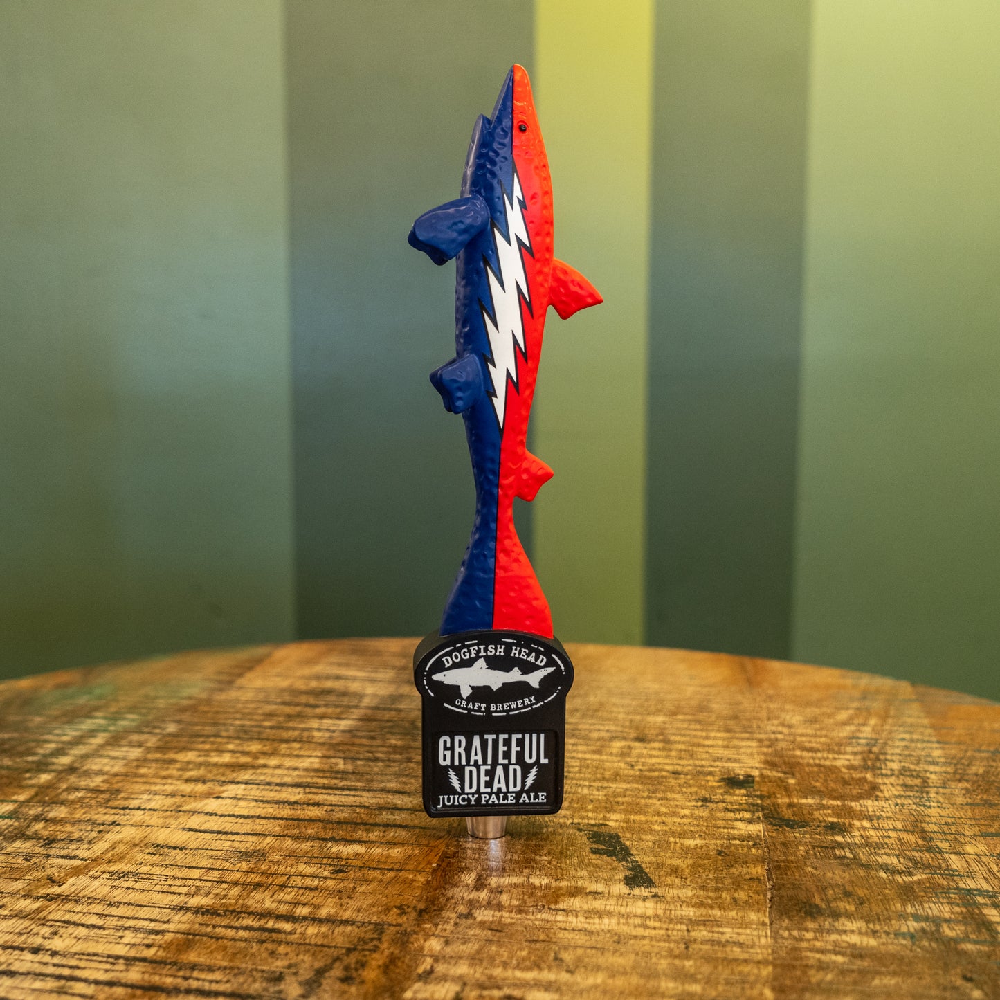 A vibrant plastic fish tap handle features a red, blue, and white lightning bolt, standing upright on a wooden table. Its base reads “Dogfish Head x Grateful Dead Tap Handle” by Merchtank, with a green striped background.