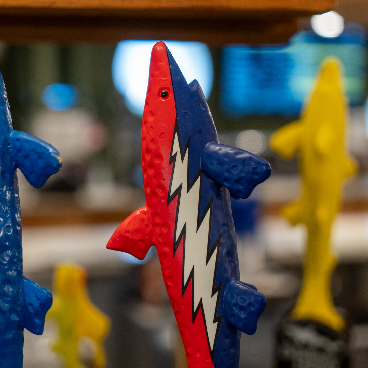The Dogfish Head x Grateful Dead Tap Handle by Merchtank features a colorful fish design with a red head, blue tail and fins, and a white zigzag lightning bolt pattern. Blurred blue and yellow fish sculptures are visible in the background.