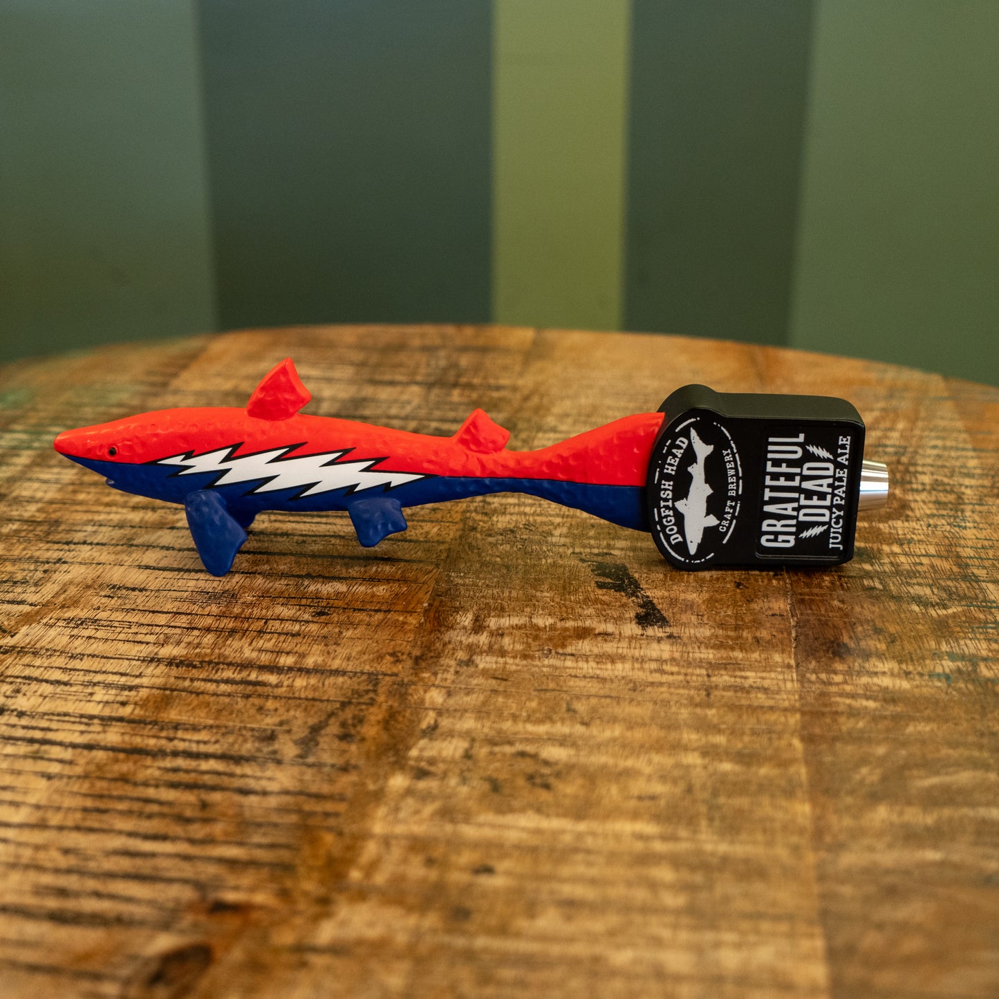A red and blue shark-shaped tap handle featuring a white lightning bolt rests on wood. It displays "Dogfish Head x Grateful Dead" with Merchtank branding on the end.
