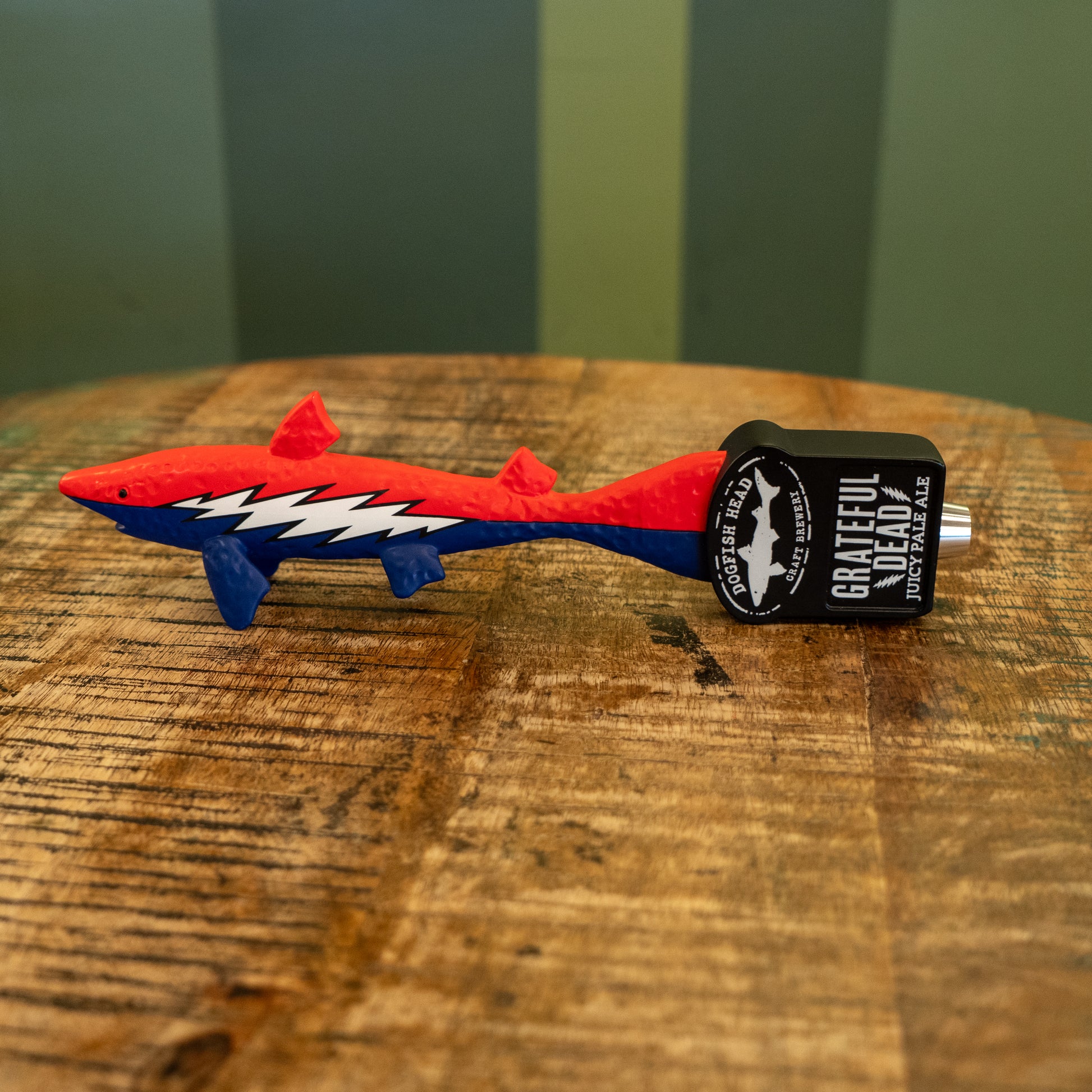 A red and blue shark-shaped tap handle featuring a white lightning bolt rests on wood. It displays "Dogfish Head x Grateful Dead" with Merchtank branding on the end.
