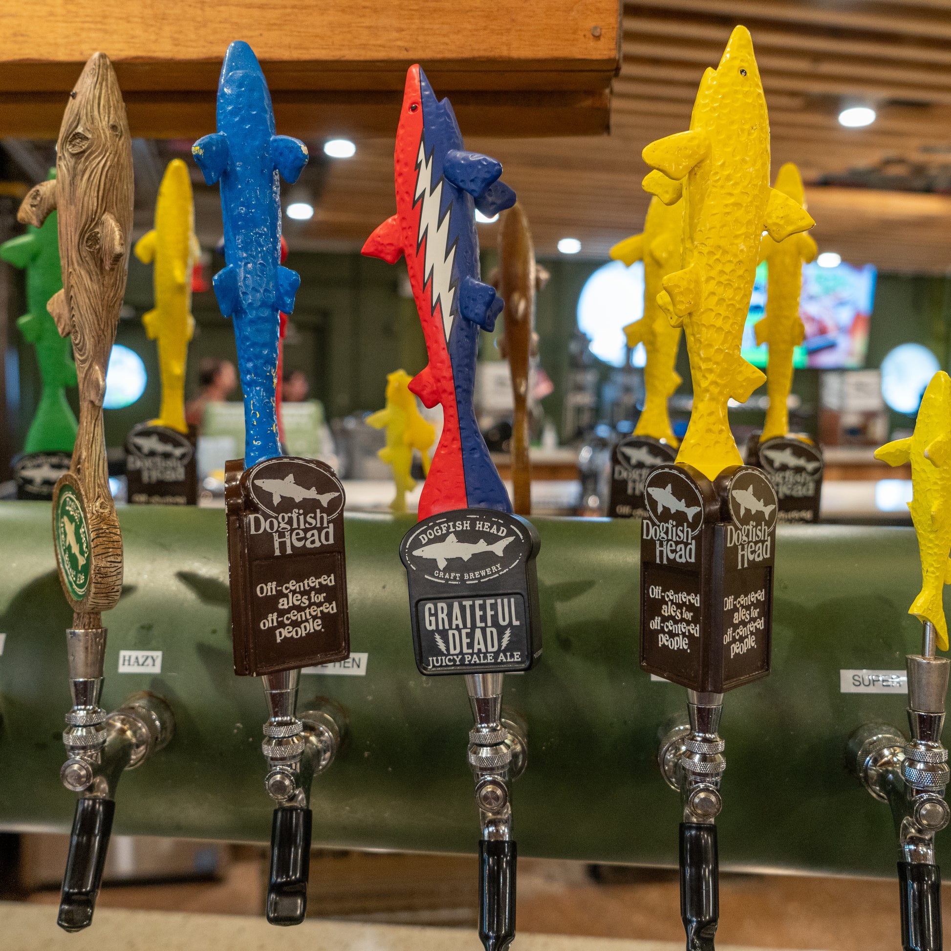 Close-up of colorful fish-shaped beer tap handles at a bar, including the Merchtank Dogfish Head x Grateful Dead Tap Handle with a red, white, and blue design featuring prominent logos and labels.