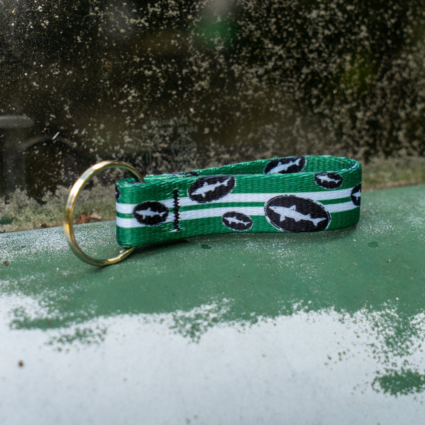 The Lupine, Inc. Green Retro Keychain features a bat logo and green, white, and black stripes—perfect for designated drivers spreading good vibes. Shown on a weathered green and white surface near a dirty window.