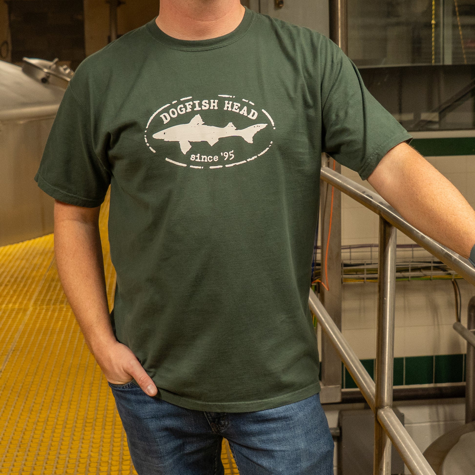 A person indoors wears the GO WEST T-SHIRT COMPANY Green Since 95 Short Sleeve Logo Tee, featuring ring-spun cotton, a fish graphic, and an oval logo celebrating "since 95," with one hand in their pocket and the other on a metal railing.