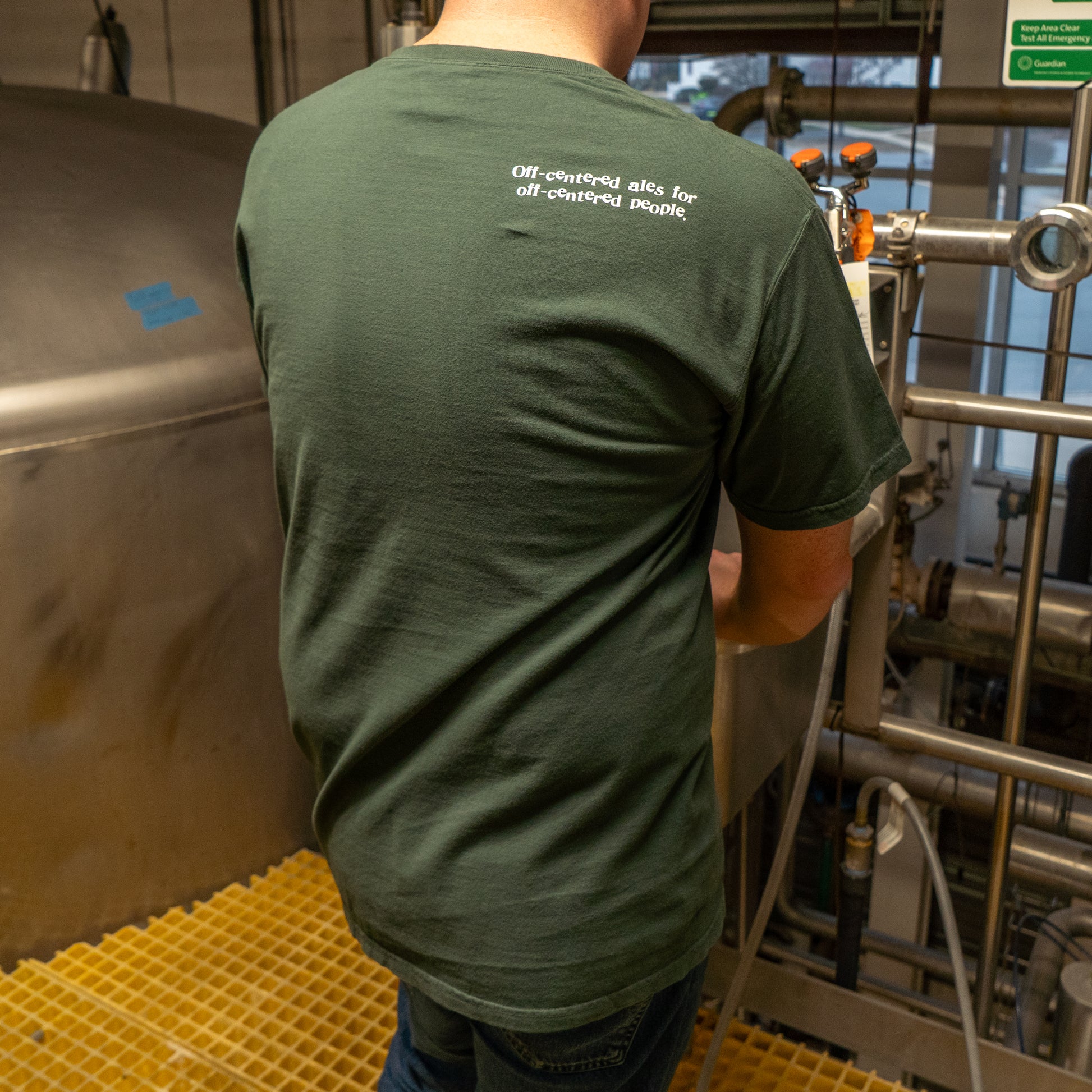 Wearing a Green Since 95 Short Sleeve Logo Tee by GO WEST T-SHIRT COMPANY, someone stands on a yellow grated floor next to stainless brewing tanks. The back says, Off-centered ales for off-centered people.