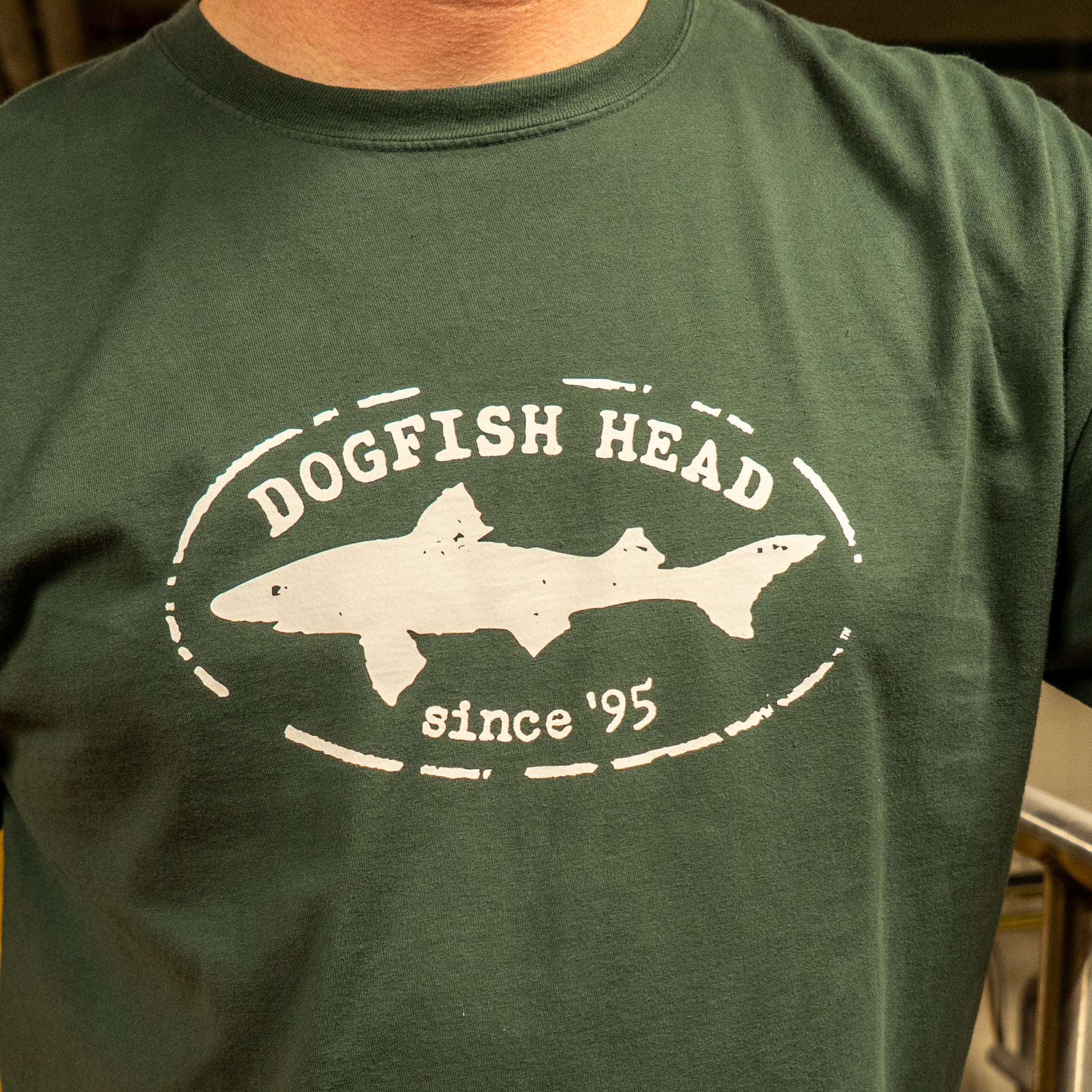 A person wearing the Green Since 95 Short Sleeve Logo Tee by GO WEST T-SHIRT COMPANY, made from soft ring-spun cotton with a white oval logo, DOGFISH HEAD text above a fish silhouette, and "since 95" below.