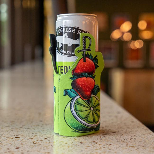 A Dogfish Head cocktail rests in a Green Swords and Lime 12oz Slim Can Cooler by Grandstand Glassware & Apparel, featuring bright green with a sliced lime, two strawberries, and a sword, set against a blurred indoor background.