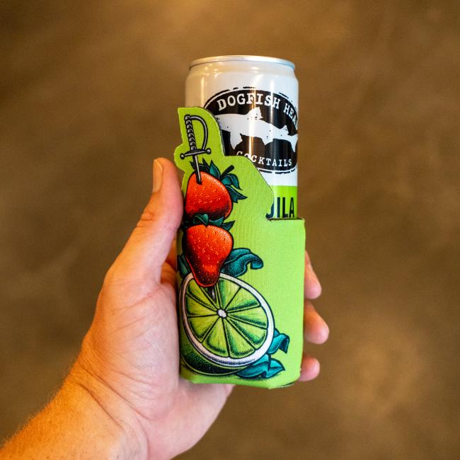 A hand holds a Dogfish Head Cocktail in the Grandstand Glassware & Apparel Green Swords and Lime 12oz Slim Can Cooler, featuring green swords, limes, strawberries, and a sword against a blurred brown background.