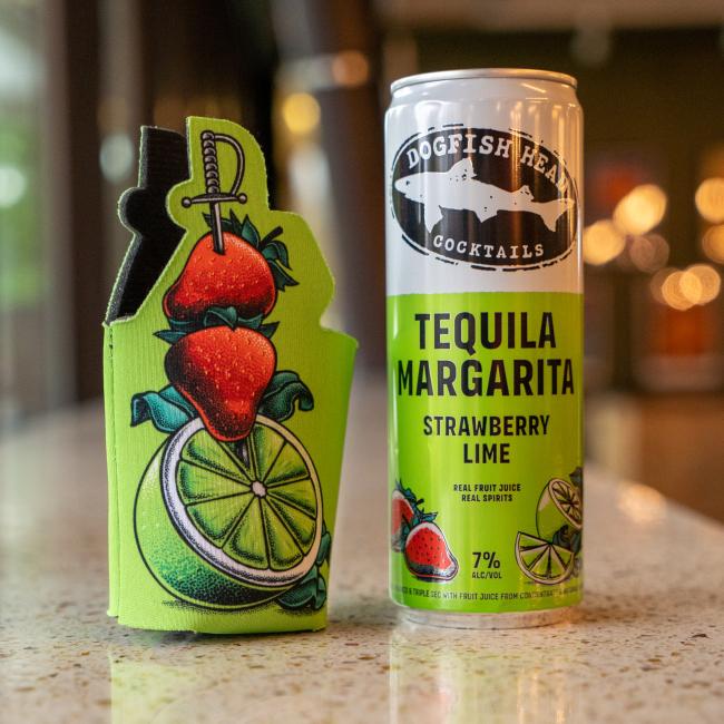 A Dogfish Head Tequila Margarita Strawberry Lime can sits beside a Grandstand Glassware & Apparel Green Swords and Lime 12oz Slim Can Cooler, with playful cartoon strawberry, lime, and dagger art. A softly blurred background enhances the scene’s vibrance.