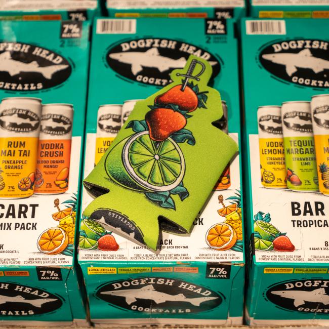 Three boxes of Dogfish Head Canned Cocktails are topped with a Grandstand Glassware & Apparel Green Swords and Lime 12oz Slim Can Cooler, featuring vibrant strawberry and lime art—perfect for keeping drinks cool in style.