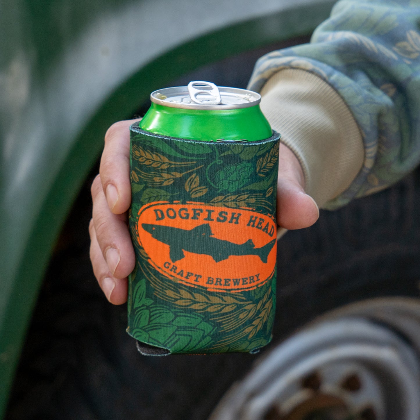 Being held with a can in it, is a green camo can cooler featuring an orange Dogfish Head shark and shield logo.