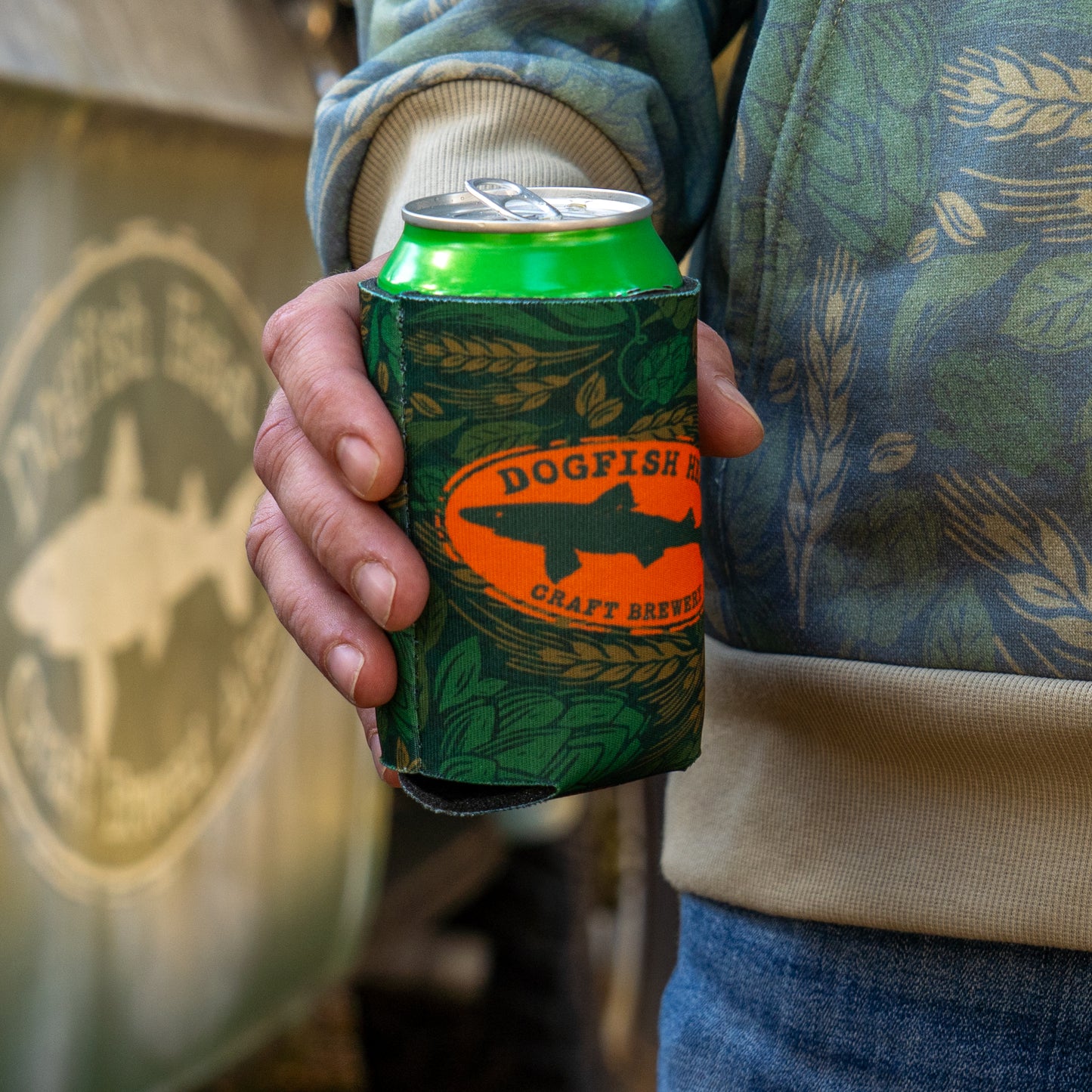 Being held with a can in it, is a green camo can cooler featuring an orange Dogfish Head shark and shield logo.